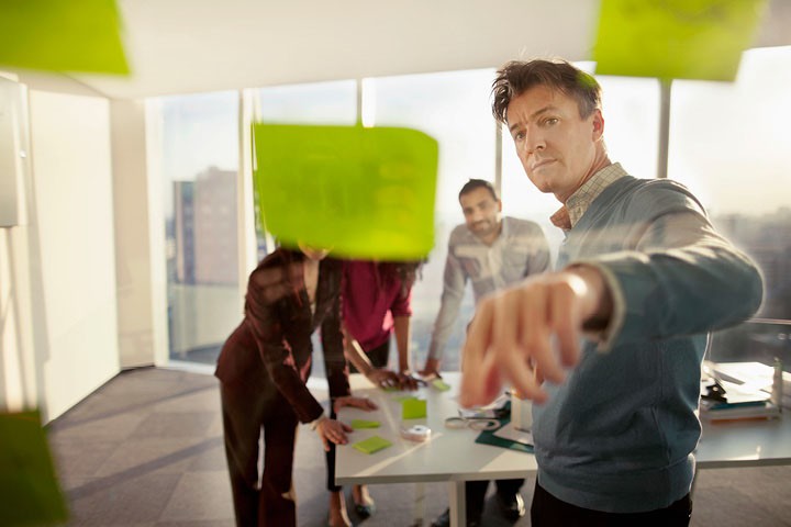 Business man with a group of people behind him in a sunny office pointing towards a transparent whiteboard
